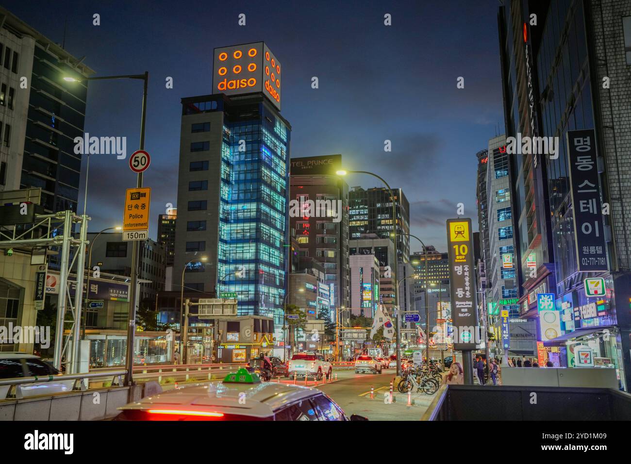 Geschäftige Stadtstraße mit einem großen Gebäude im Hintergrund. Es gibt viele Autos und Motorräder auf der Straße Stockfoto