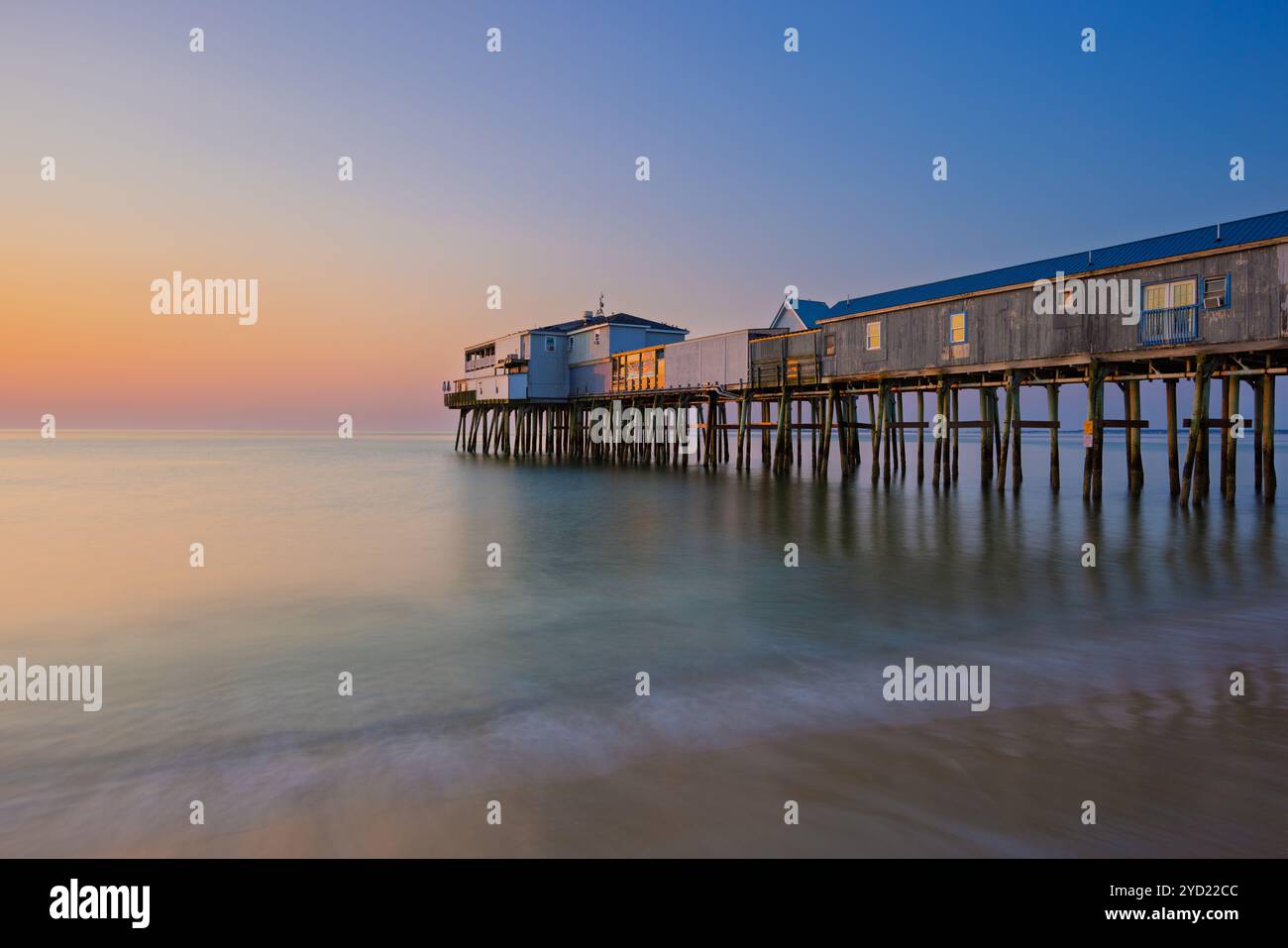 Eine ruhige Ozeanszene bietet einen langen Holzpier, der sich in ruhiges Wasser erstreckt, mit einem weichen Farbverlauf am Sonnenuntergangshimmel. Die ruhige Umgebung ist Stockfoto