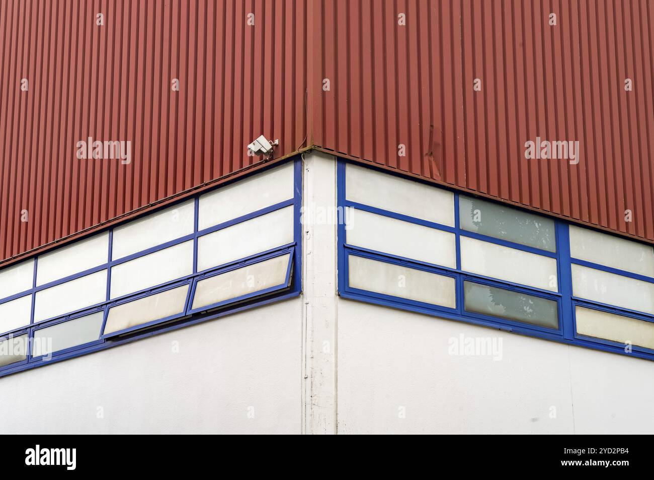 An der Ecke eines roten Industriegebäudes mit blau gerahmten Fenstern befindet sich eine einzige Überwachungskamera Stockfoto