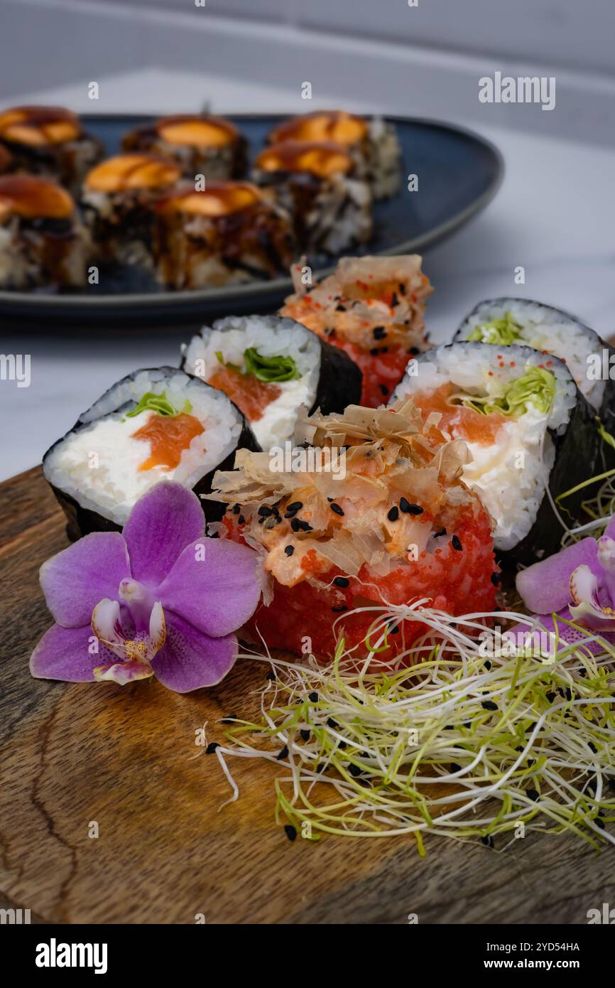 Frische Sushi-Brötchen mit Lachs, Frischkäse und Sprossen werden auf einem Holzbrett serviert, das mit Orchideenblüten dekoriert ist Stockfoto