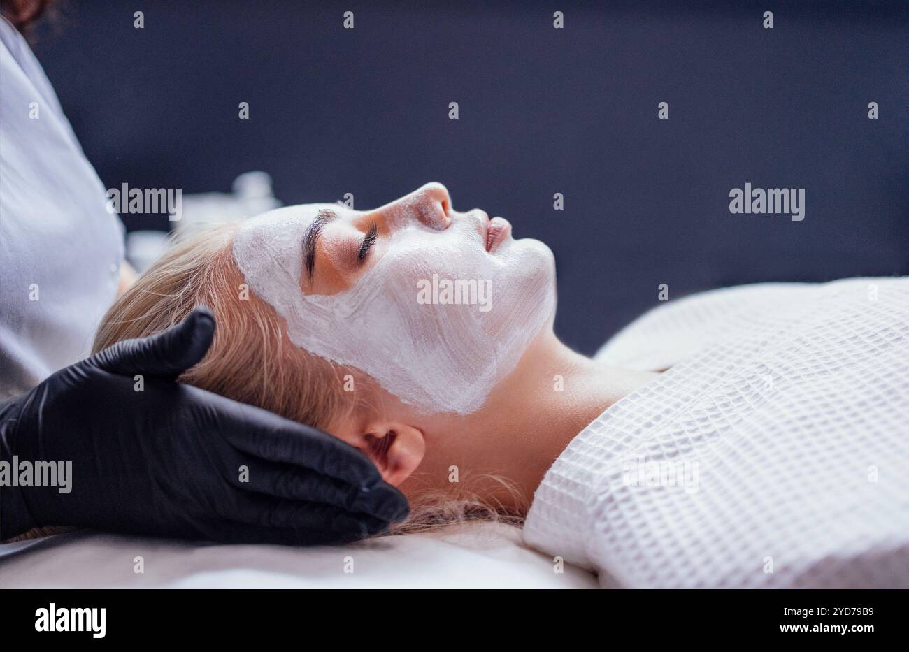 Der Kosmetiker trägt eine verjüngende weiße Maske auf das weibliche Klientelgesicht auf Stockfoto