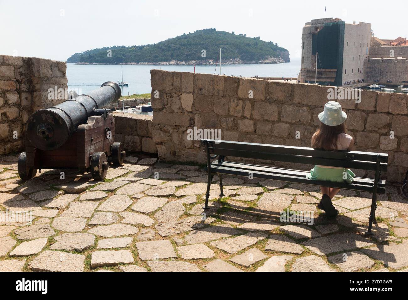 Junge Mädchen / Kind / Kind genießen die Aussicht vom Aussichtspunkt an der Seite von Fort Revelin, nach dem Ploce Gate Eingang in die ummauerte Altstadt. Dubrovnik. Kroatien. Die Bank ist schlecht platziert, da die Sicht durch die Verteidigungsmauer eingeschränkt ist. Eine originale Kanonenartillerie-Pistole ist zu sehen. (138) Stockfoto