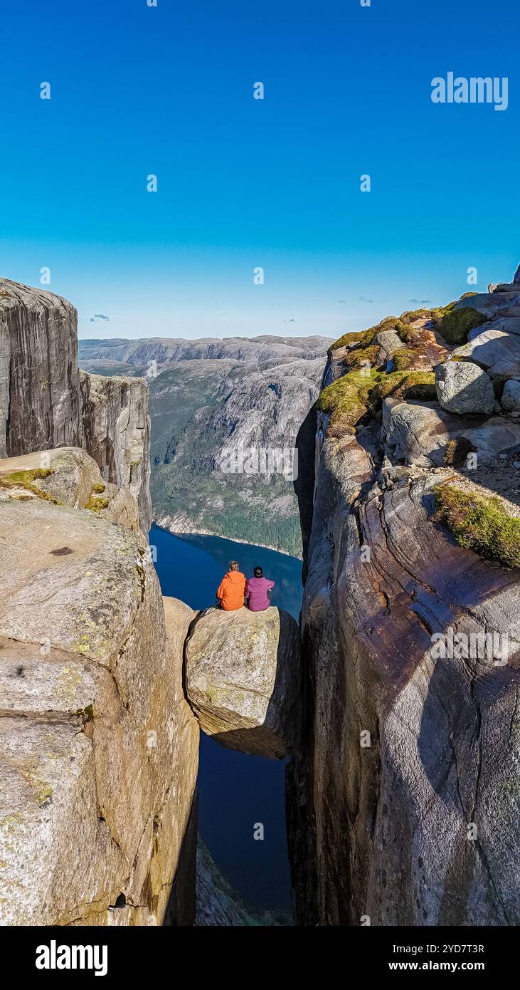 Zwei Individuen sitzen auf einem schmalen Felsvorsprung mit Blick auf ein tiefes Tal in der NorwaynKjeragbolten Klippe Stockfoto
