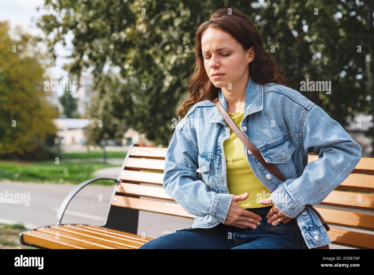 Frau in lässiger Kleidung, die auf einer Bank sitzt und einen Teil ihres Bauches mit beiden Händen aufgrund von Schmerzen hält. Stockfoto