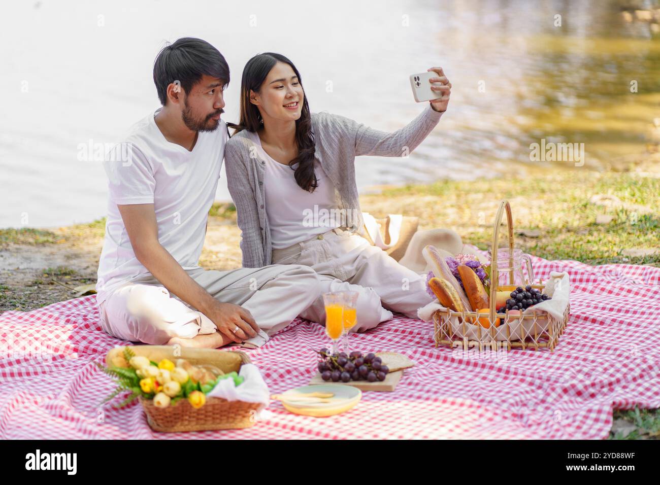 Verliebtes asiatisches Paar, das Selfie mit Smartphone macht, entspannendes togetherÂ mit Picknickkorb Stockfoto