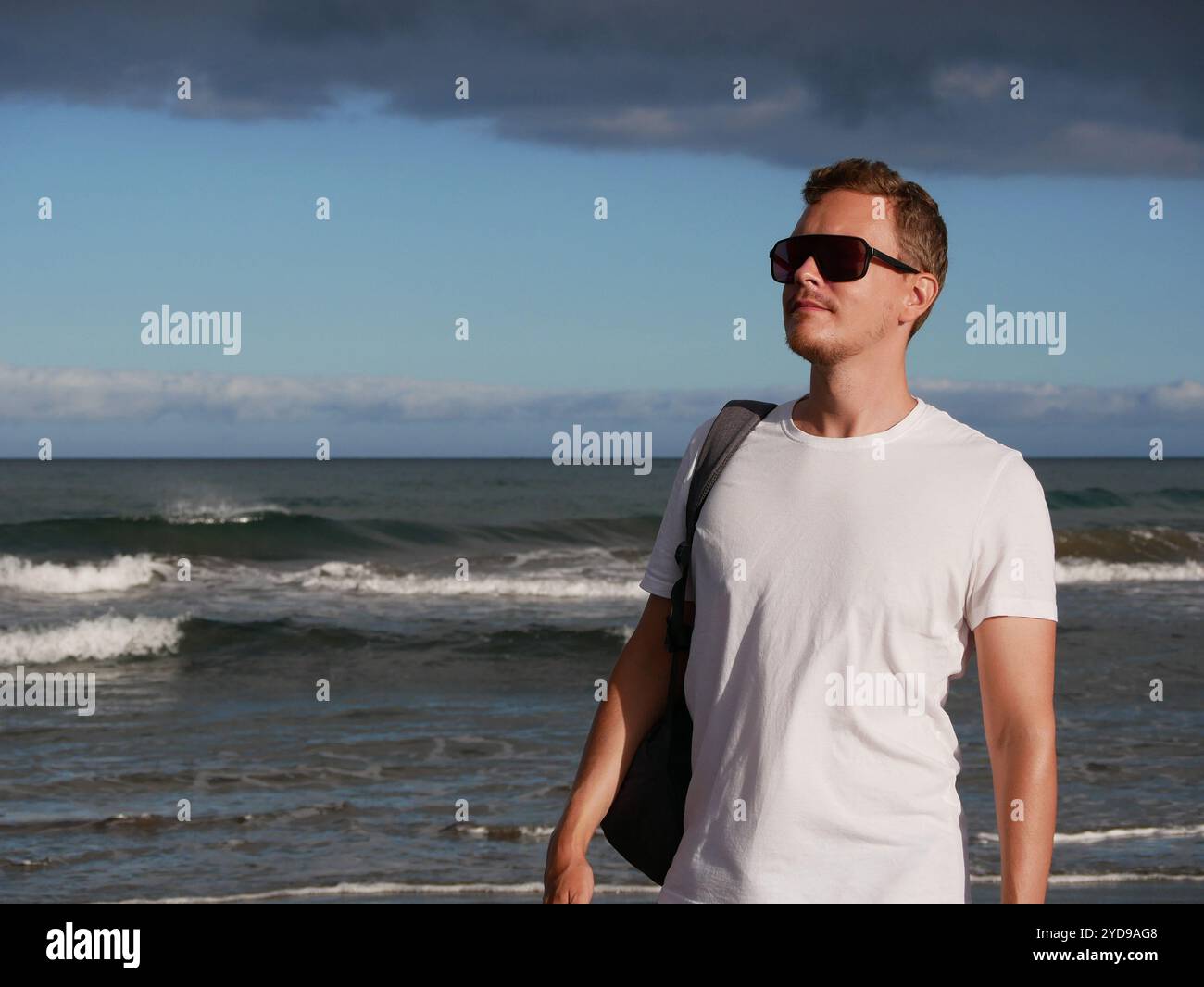 Ein junger Mann mit Rucksack, mit weißem T-Shirt und Sonnenbrille, der am Meer vor dem Hintergrund des trüben Wolkenhimmels steht, mit Kopierraum. Stockfoto