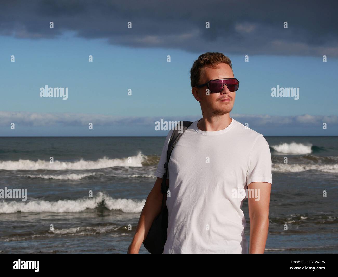 Ein junger Mann mit Rucksack, mit weißem T-Shirt und Sonnenbrille, der am Meer vor dem Hintergrund des trüben Wolkenhimmels steht, mit Kopierraum. Stockfoto