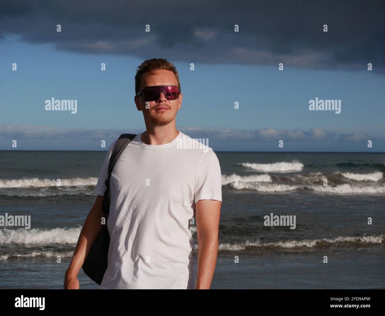 Ein junger Mann mit Rucksack, mit weißem T-Shirt und Sonnenbrille, der am Meer vor dem Hintergrund des trüben Wolkenhimmels steht, mit Kopierraum. Stockfoto