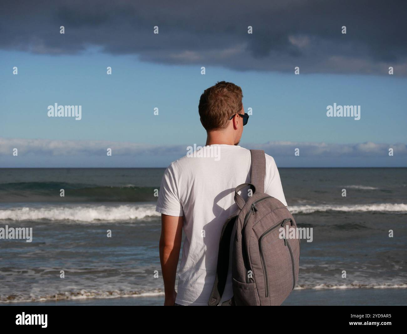 Ein junger Mann mit Rucksack, mit weißem T-Shirt und Sonnenbrille, der auf das Meer blickt, vor dem Hintergrund des trüben, bewölkten Himmels, mit Kopierraum. Touris Stockfoto
