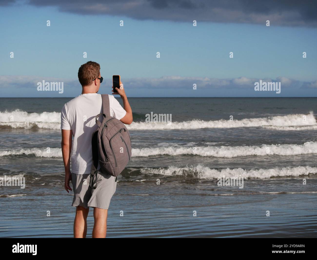 Ein junger Mann mit Rucksack, mit weißem T-Shirt und Sonnenbrille, der ein Foto vom Meer vor dem Hintergrund des trüben bewölkten Himmels macht, mit Kopierraum. Bis Stockfoto