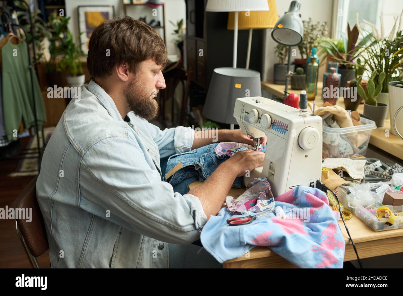 Bärtiger Mann, der ein Stück Textil zu einer Second Hand Denim Jacke auf einer elektrischen Nähmaschine näht, während er gebrauchte Kleidung aufrüttelt Stockfoto