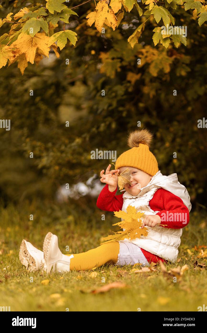 Ein lächelndes Kind in einem hellen Outfit sitzt auf dem Gras und spielt fröhlich mit lebhaften Herbstblättern, umgeben von bunten Blättern an einem sonnigen Tag. Stockfoto
