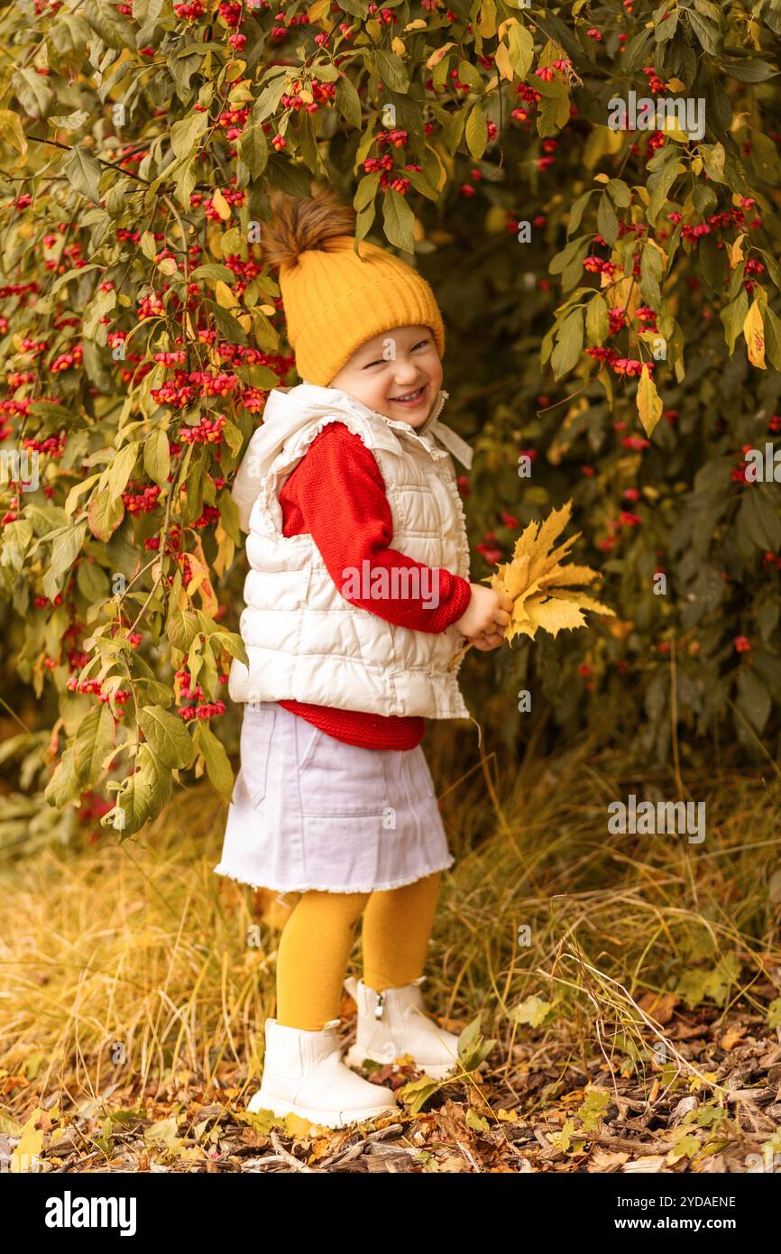 Ein fröhliches Kind steht inmitten lebendiger Blätter und trägt eine gelbe Mütze und eine gemütliche Weste. Sie lächelt, während sie bunte Herbstblätter hält, umgeben von Stockfoto