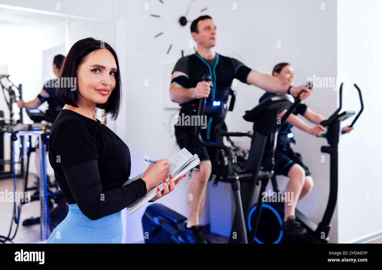 Die junge, lächelnde Trainerin zeichnet während des Trainings ihre Kundendaten auf. Ein Mann im ems-Anzug trainiert auf einem Ellipsentrainer. Frau Stockfoto