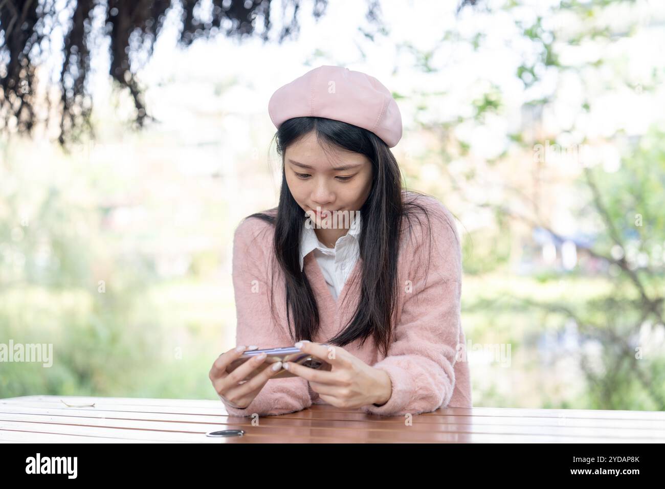 Eine langhaarige taiwanesische Frau im Alter von 20 Jahren, die einen rosafarbenen Pullover trägt, bedient ein Smartphone, während sie auf einer Parkbank im Bezirk Xinyi, Taipei, Ta, sitzt Stockfoto