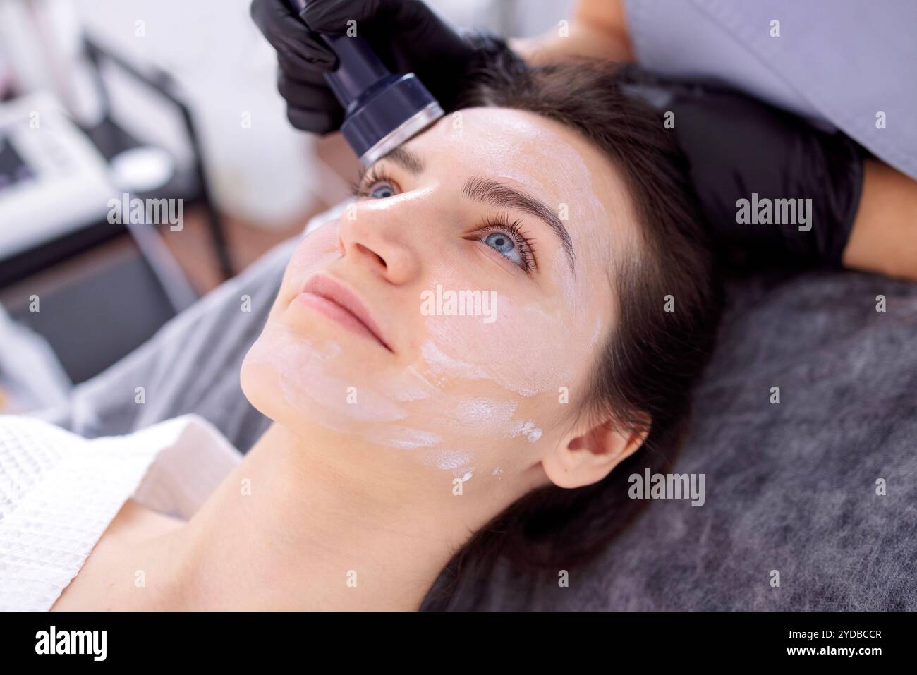Eine junge Frau im Bademantel liegt und erhält mit einem Gerät eine Gesichtsreinigung Stockfoto