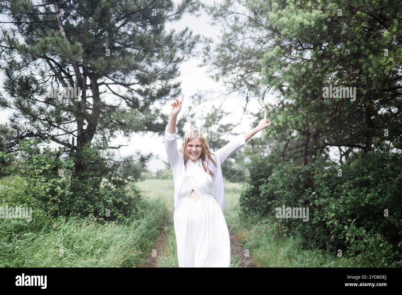 Porträt einer lachenden jungen Frau in einem weißen Kleid im Regen. Eine nasse Blondine hat Spaß und tanzt im Regen im Park. Stockfoto