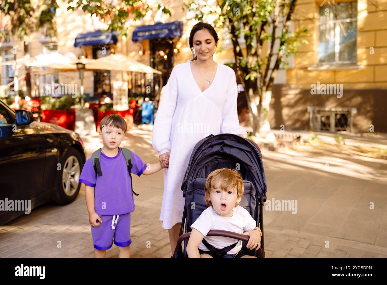 Schwangere Frau in weißem Kleid spaziert mit ihren beiden Kindern durch die Stadt. Mutter mit Kinderwagen führt ihr ältestes Kind von Hand auf die Straße Stockfoto