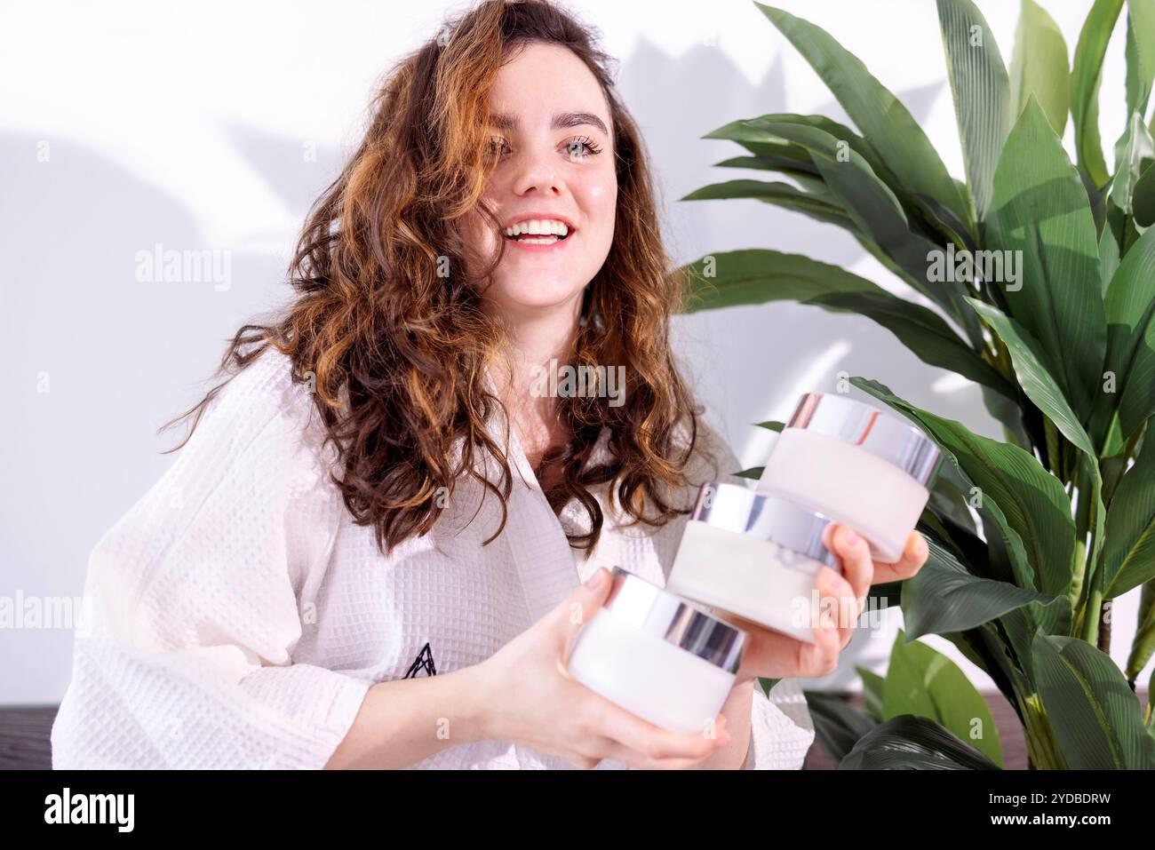 Nahaufnahme eines lockigen, attraktiven Mädchens, das ein Modell aus Cremedosen in einem Spa hält. Eine junge Frau im Bademantel lacht und wählt Stockfoto