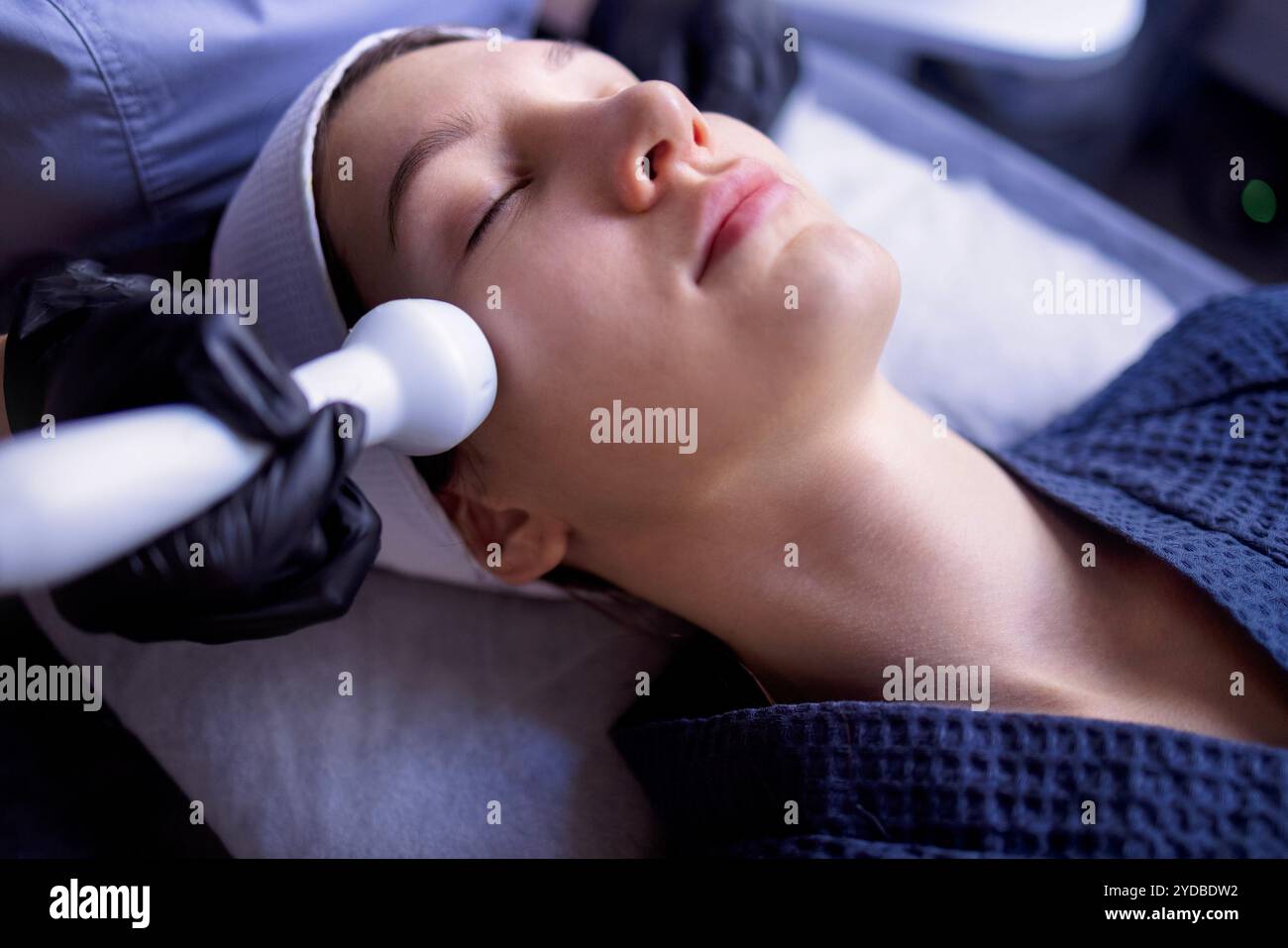 Eine junge Frau im Bademantel liegt und erhält eine Gesichtsreinigung mit einem Gerät in einem Spa- oder Schönheitszentrum. Ein natürliches f Stockfoto