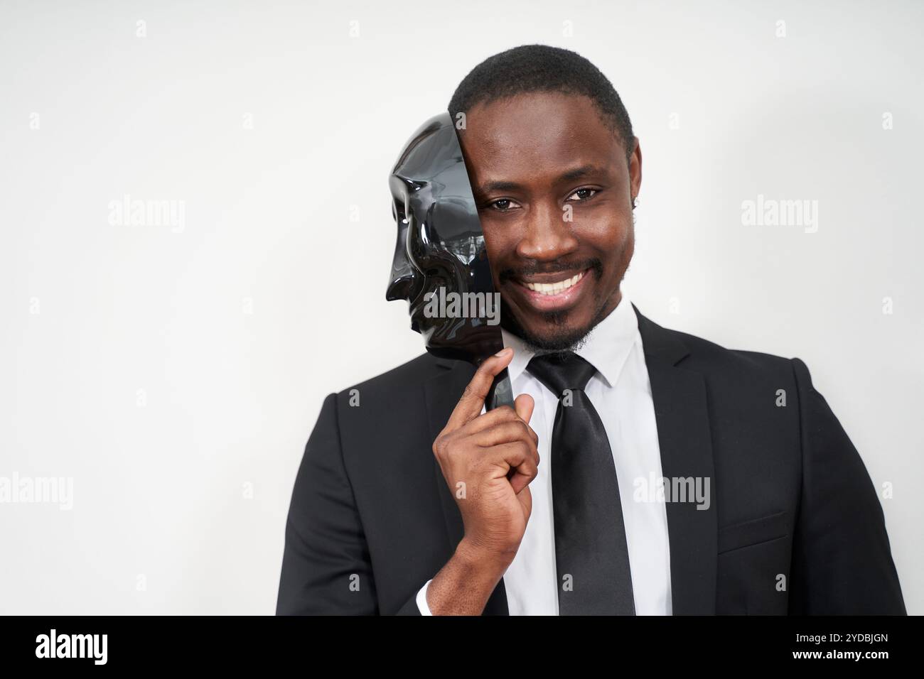 Afrikanischer junger Mann mit schwarzem Anzug, der eine Plastikmaske abnimmt, die das Gesicht enthüllt und lächelt Stockfoto