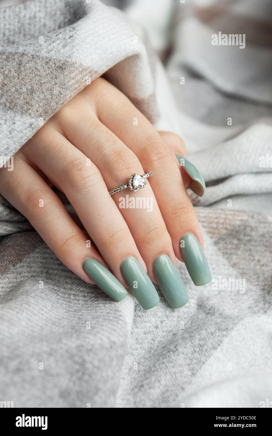 Eine Hand zeigt lange, elegant gepflegte Nägel, die in einem weichen Grünton lackiert sind. Die Hand liegt auf einer gemütlichen, gemusterten Decke, die ein funkelndes Licht hervorhebt Stockfoto