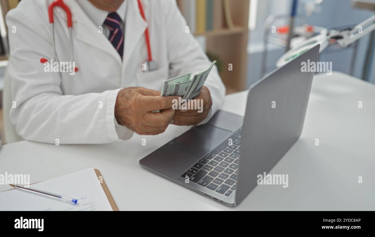 Senior männlicher Arzt in einem weißen Mantel, der uns Dollar an seinem Schreibtisch mit einem Laptop in einem Krankenhauszimmer zählt. Stockfoto