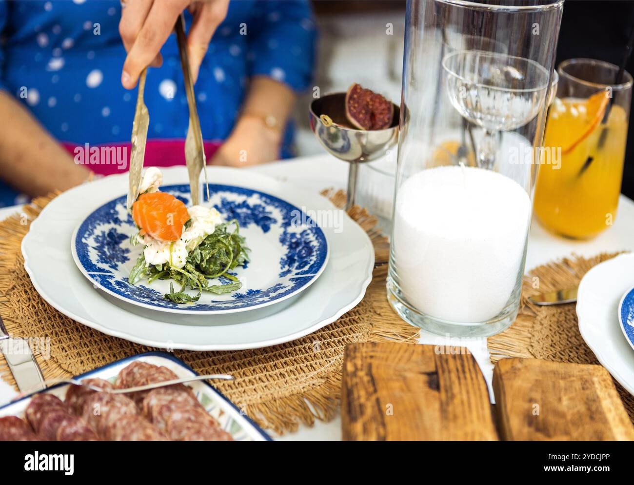 Ein Kellner serviert frischen Lachssalat auf einem blau dekorierten Teller für einen Gast. Der Tisch ist mit Kerzen und gewebten Tischsets dekoriert. Brot, Fleisch, drin Stockfoto