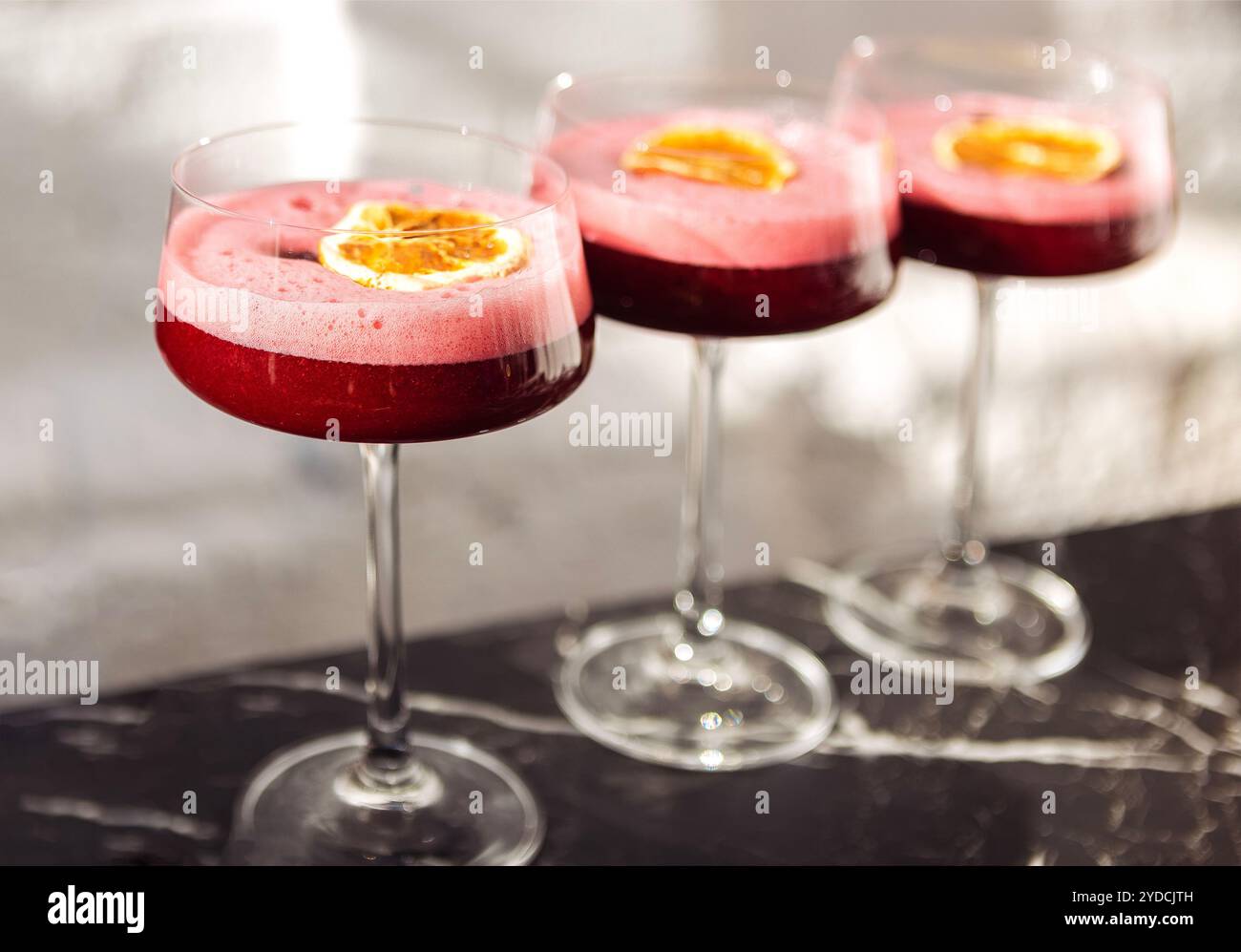 Rosafarbene süß-saure Beerencocktails mit flauschigem Schaum in eleganten, langstieligen Gläsern. Köstliche Getränke sind mit einem Stück Orange und Blumen dekoriert Stockfoto