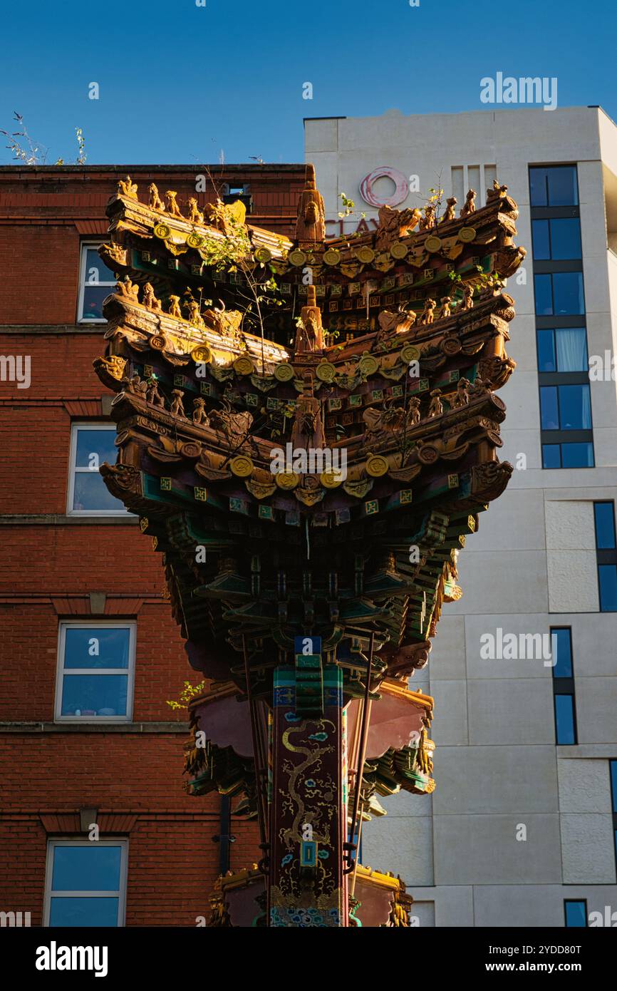 Vor einem weißen, modernen Gebäude steht eine farbenfrohe, aufwändig geschnitzte Dachkonstruktion mit goldenen, grünen und roten Details Stockfoto