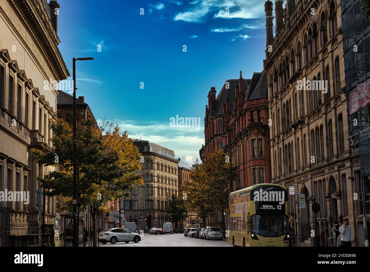Eine Stadtstraße mit hohen Backsteinhäusern, einem gelben Doppeldeckerbus und Autos, die am Straßenrand geparkt sind. Der Himmel ist blau mit weißen Wolken im Menschen Stockfoto