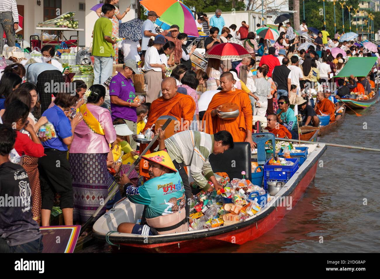 Zwei Mönche, die auf einem Ruderboot stehen und gesehen werden, wie sie Almosen von Menschen empfangen, die am Ufer des Chao Phraya am Wat Chin Wararam Worawihan Tempel in der Provinz Pathum Thani, Thailand, für sie anstehen. Die Tradition, hundert Mönchen in Pathum Thani, Thailand, Almosen zu geben, ist eine jahrhundertealte buddhistische Zeremonie, die entlang von Flüssen und Kanälen nach dem Ende der buddhistischen Fastenzeit stattfindet. Mönche versammeln sich in Booten, um Opfer von Einheimischen zu erhalten, die Essen zubereiten und an der Verdienste teilnehmen. Diese Veranstaltung feiert Gemeinschaft, Glauben und kulturelles Erbe. Stockfoto