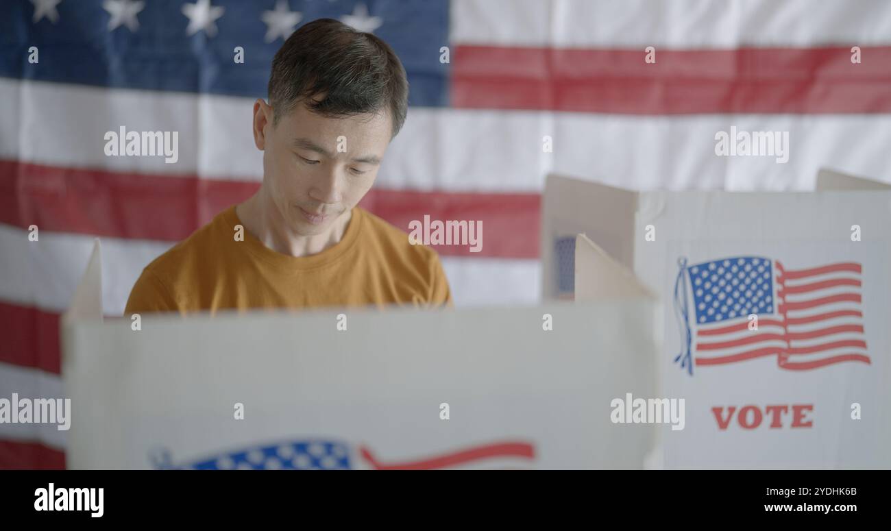 Frontale Nahaufnahme des Asiaten, der Stimmen in der amerikanischen Wahlkabine abgibt. Leerer Stand und US-Flagge im Hintergrund Stockfoto