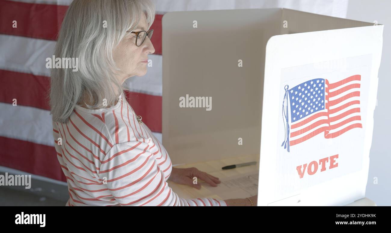 Hoher Blickwinkel über der Schulter, Seniorin, die allein am Stand stimmte, mit US-Flagge an der Wand dahinter. Haare unten, Brille Stockfoto