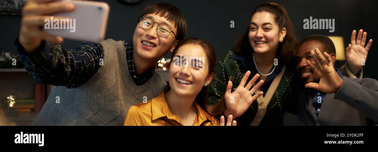 Freimütige Aufnahme eines asiatischen Mannes, der ein Selfie mit vier fröhlichen Freunden macht, die Hände vor der Kamera winken, während er am festlichen Esstisch sitzt und Weihnachten zu Hause feiert Stockfoto