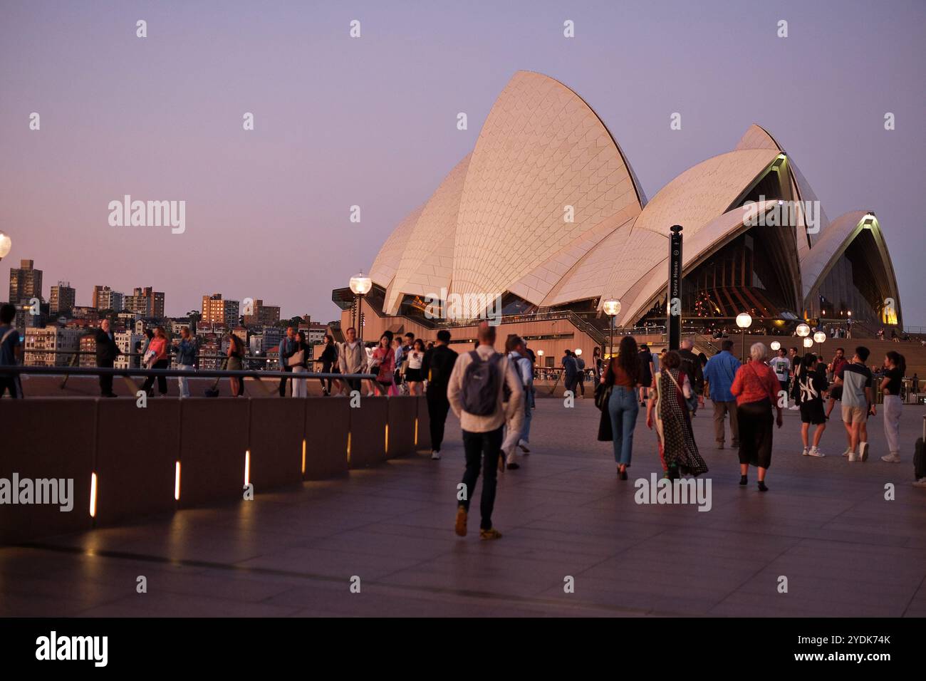 Wir nähern uns dem Opernhaus von Sydney in der Abenddämmerung mit Menschen, die auf dem Vorplatz spazieren gehen, die Segel in sanftem Licht, Gebäude von Kirribilli in der Ferne Stockfoto