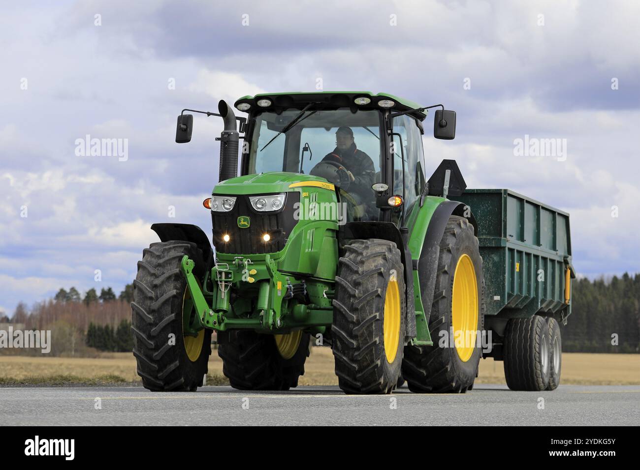 Landwirt fährt John Deere 6150R-Traktor und landwirtschaftlichen Anhänger auf der Straße. 2018 verzögerte sich die Aussaat im späten Frühjahr. Jokioinen, Finnland, 30. April 2018, Euro Stockfoto