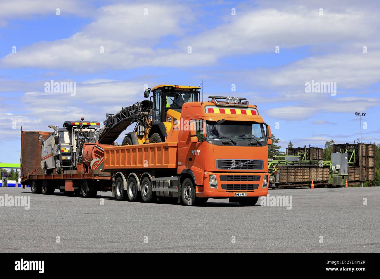 Orange Volvo FH transportiert an einem Sommertag mit blauem Himmel und Wolken Baumaschinen auf einem Anhänger auf einem Asphaltplatz. Orivesi, Finnland, 14. Juni 2018 Stockfoto