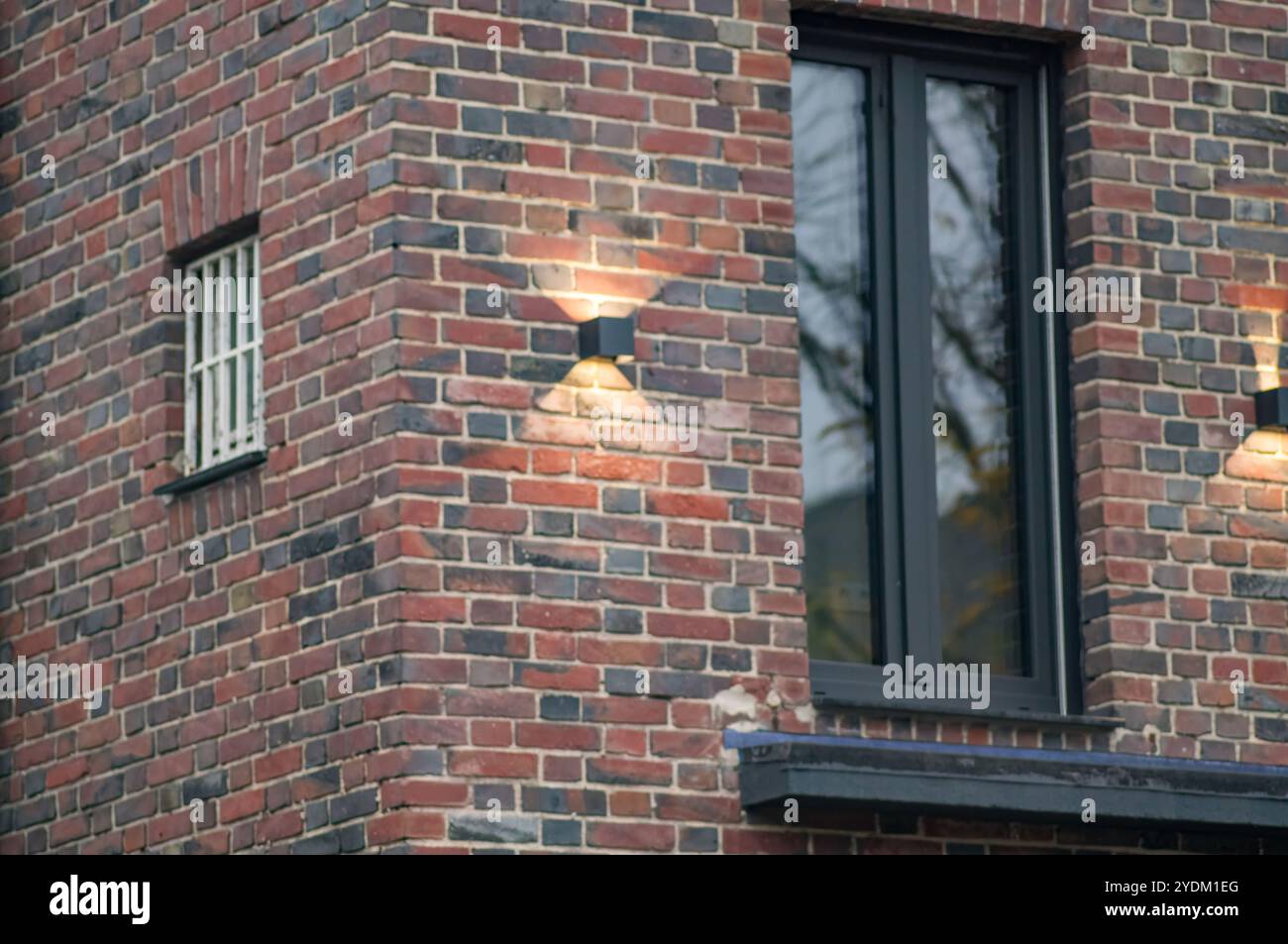 Außenwandleuchte beleuchtet eine rote Backsteinwand eines modernen Gebäudes mit einem Fenster und einem vergitterten Kellerfenster Stockfoto