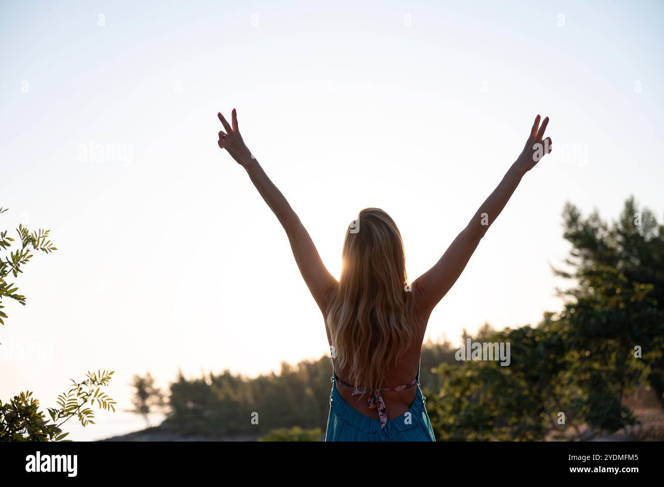 Eine Frau mit ausgestreckten Armen blitzt Friedenszeichen vor einem glühenden Sonnenuntergang, umgeben von der Natur, symbolisiert Freude, Freiheit und Frieden. Stockfoto