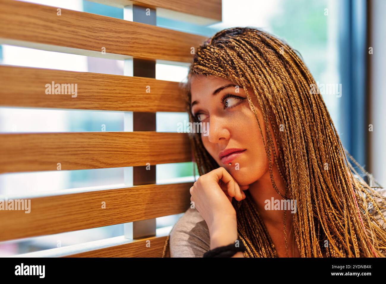 Eine junge Frau mit umwerfenden Flechten lehnt sich an eine rustikale Holzwand. In der Nachdenklichkeit verloren. Ihre Hand liegt auf ihrem Kinn, während sie nachdenklich schaut Stockfoto