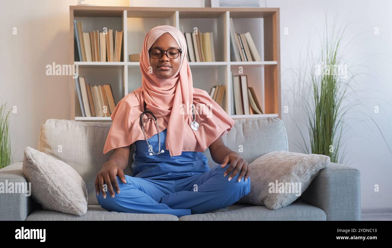 Arbeitsmeditation. Entspannender Arzt. Yoga Freizeit. Achtsame ruhige Frau Krankenschwester, die Stressentlastungsübungen in Lotusstellung auf der Couch im Krankenhaus praktiziert. Stockfoto