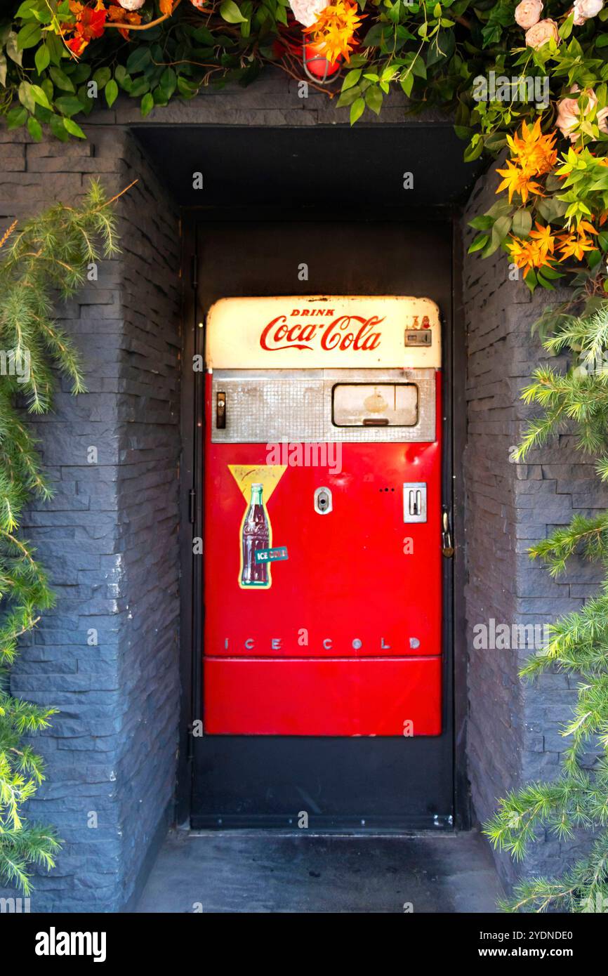 Roter Coca-Cola-Automat in einem Steineingang, umgeben von üppigem Grün und lebendigen Blumen Stockfoto