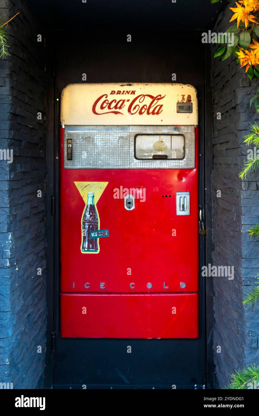 Roter Coca-Cola-Automat in einem Steineingang, umgeben von üppigem Grün und lebendigen Blumen Stockfoto