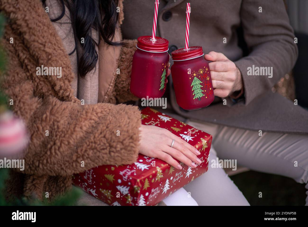 Ein Paar teilt sich einen Moment der Freude während der Weihnachtszeit und hält festliche Getränke, während es neben einem wunderschön geschmückten Weihnachtsbaum und einem Wr sitzt Stockfoto