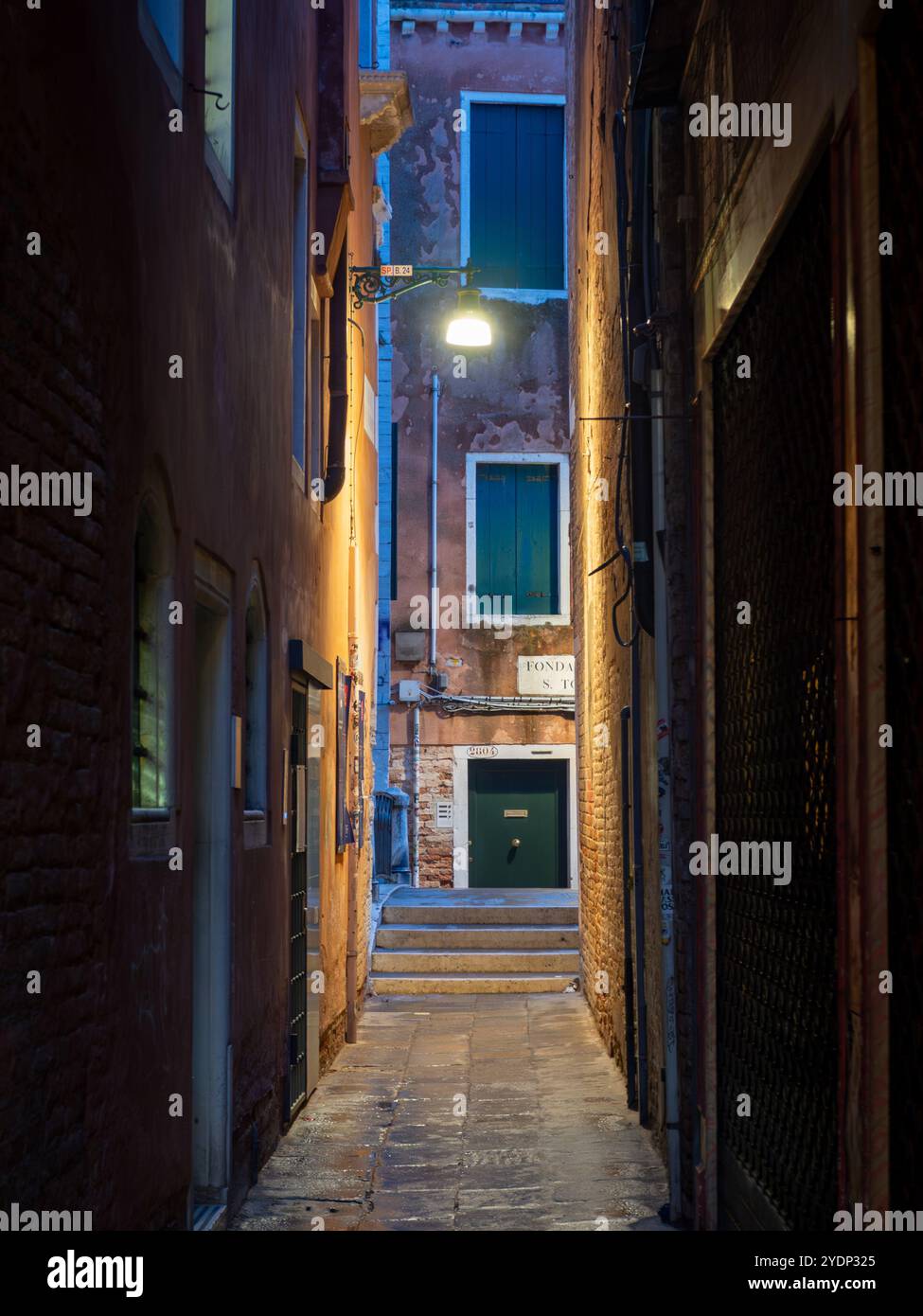 Enge Gassen in Venedig, beleuchtet von einer warmen Straßenlaterne, während der nasse Kopfsteinpflasterboden das Stadtbild über den Gebäuden reflektiert. Stockfoto