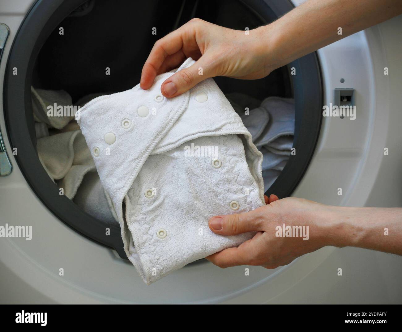 Weibliche Hände halten eine Babywindel aus weißem Tuch vor einer Waschmaschine voller waschbarer Bambuswindeln. Umweltfreundlicher wiederverwendbarer Stoff nappi Stockfoto