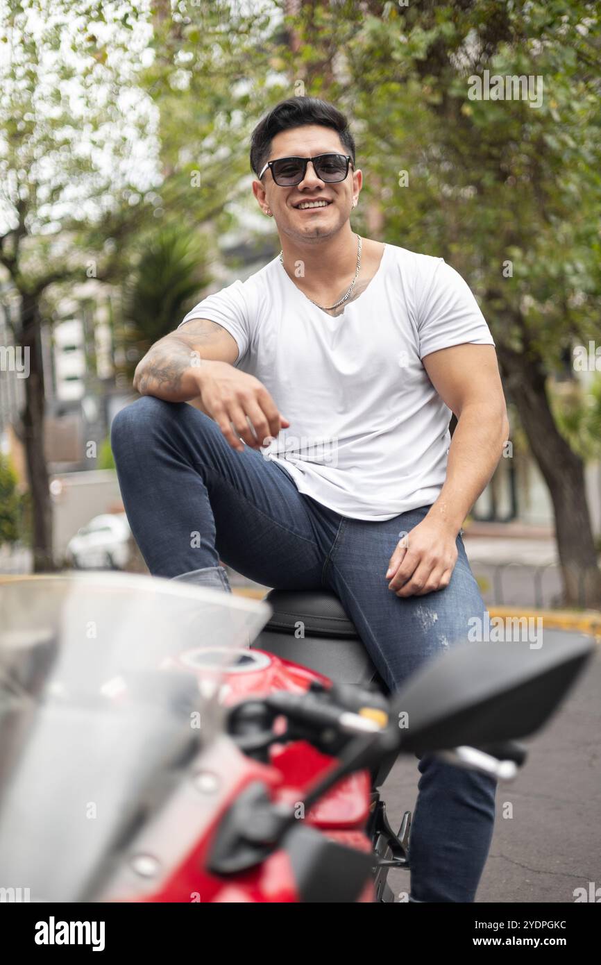 Fröhlicher junger Mann mit kurzen Haaren, lässige Kleidung und Sonnenbrille, männliches Model sitzt an einem sonnigen Tag auf seinem Motorrad, Lebensstil, Person Stockfoto