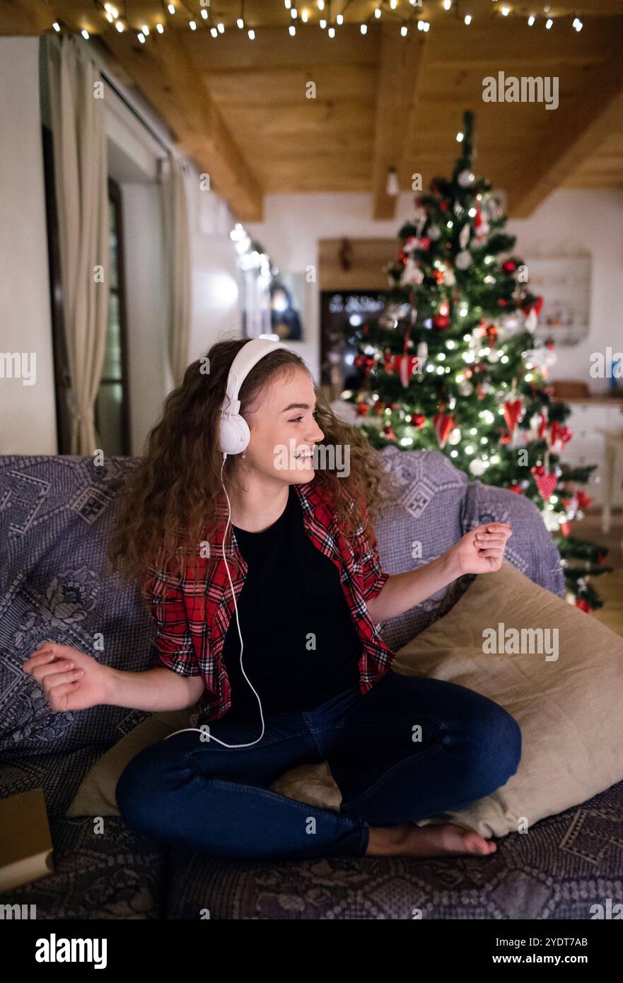 Ein Student, der Hygge Weihnachten zu Hause hat. Die Frau sitzt auf dem Sofa am Weihnachtsbaum und hört Musik mit Kopfhörern. Stockfoto