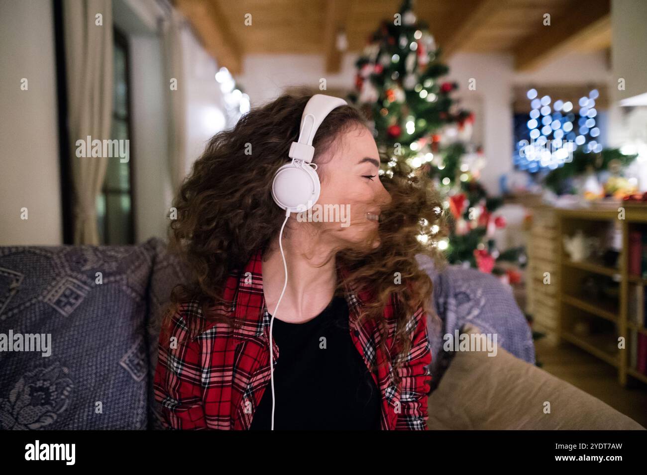 Ein Student, der Hygge Weihnachten zu Hause hat. Die Frau sitzt auf dem Sofa am Weihnachtsbaum und hört Musik mit Kopfhörern. Stockfoto
